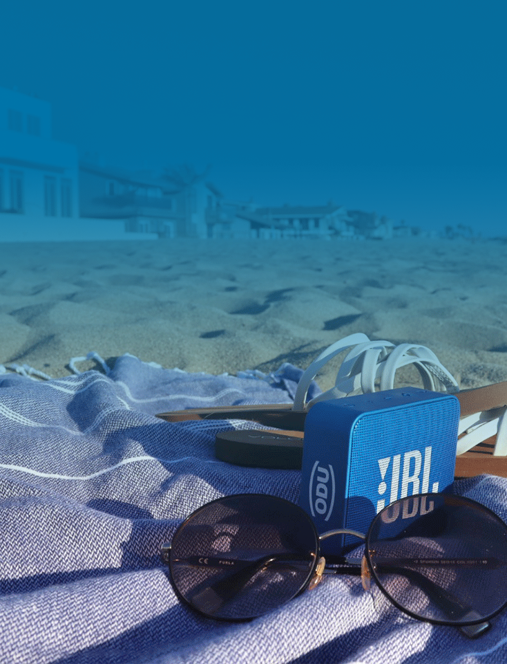 Sunglasses, blue ODU music box and sandals on a blanket on the beach.